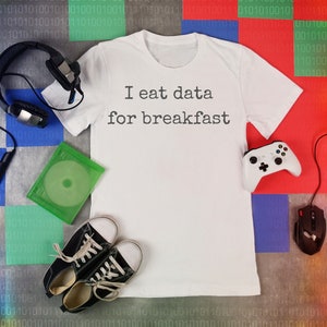 May include: A white t-shirt with the text "I eat data for breakfast" printed on it. The shirt is surrounded by gaming accessories, including a headset, a game controller, a mouse, and a game disc.