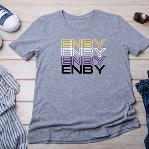 May include: A gray t-shirt with the word "ENBY" repeated four times in black, yellow, purple, and white lettering. The shirt is laid flat on a wooden surface with a pair of blue jeans, a blue and white striped shirt, and a pair of blue sneakers.