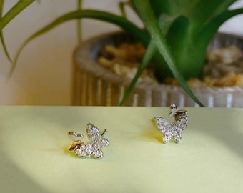 May include: A pair of silver butterfly earrings with clear crystal accents. The earrings are on a light green surface.