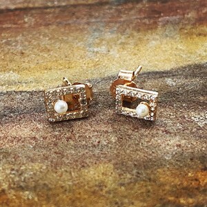 May include: A pair of gold-colored square stud earrings. Each earring features a small, round pearl in the center, surrounded by a square frame of small, clear crystals. The earrings are set against a textured, brown surface.