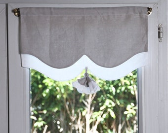Puede incluir: Una cenefa gris y blanca con forma de concha con un borla de tela blanca que cuelga del centro. La cenefa está colgada en una ventana con vista a un follaje verde.