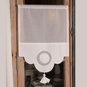 Puede incluir: Cortina blanca de voile con un borde inferior festoneado y un círculo bordado decorativo con un diseño floral. La cortina tiene un borla blanca.