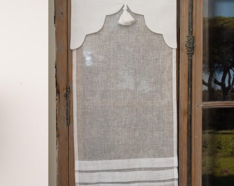 May include: A white linen window valance with a scalloped top and a decorative tassel. The valance hangs over a sheer, light-colored curtain with horizontal stripes at the bottom. The window frame is made of dark wood.