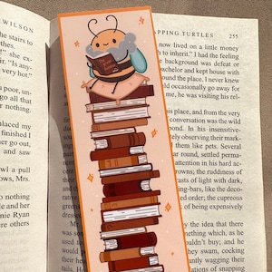 Könnte beinhalten: Ein skurriles Lesezeichen mit einer Cartoon-Biene, die "The Great GatBEE" auf einem Stapel Bücher liest. Das Lesezeichen hat einen orangefarbenen Rand und Sternakzente. Die Bücher sind in Brauntönen gehalten.