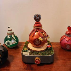May include: A decorative, reddish-brown bottle with a heart-shaped stopper and gold chain. The bottle sits on a mossy base with a wooden slice and miniature mushrooms. The word "AURORA" is visible on the bottle.