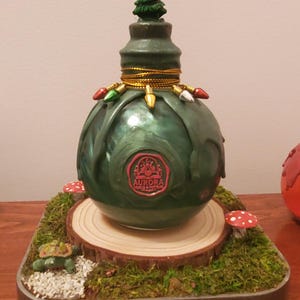 May include: A decorative Christmas-themed ornament with a green, bulbous body and a small Christmas tree on top. The ornament is adorned with string lights and rests on a base with moss, mushrooms, and a wooden slice. The word "AURORA" is visible.