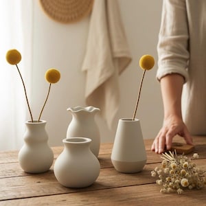 Peut inclure: Quatre vases en céramique blanc cassé de formes variées sont disposés sur une surface en bois. Deux vases contiennent des fleurs sphériques jaunes. Les vases ont une finition mate et des hauteurs et designs différents.