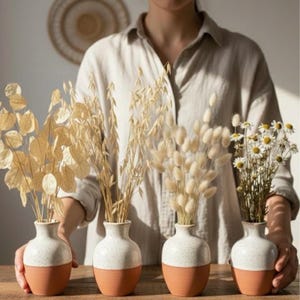 Könnte beinhalten: Vier kleine Keramikvasen mit Trockenblumen. Die Vasen haben eine gesprenkelte weiße obere Hälfte und einen terrakottafarbenen Sockel. Die Trockenblumen umfassen Silberblatt, Hafer, Hasenschwänze und Gänseblümchen.