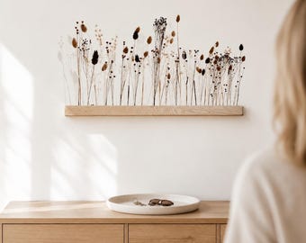 Puede incluir: Un estante de pared rectangular de madera clara sostiene un arreglo de hierbas y flores secas en tonos beige, marrón y negro. Los tallos están erguidos y tienen alturas variadas, creando una exhibición lineal de elementos botánicos.