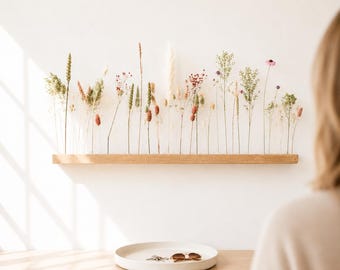 Puede incluir: Un estante rectangular de madera clara montado en la pared, que sostiene un arreglo de varias hierbas secas, flores silvestres y tallos en tonos verde, beige, rosa y blanco. Debajo del estante hay una bandeja redonda y poco profunda de cerámica blanca.