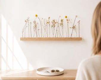 Puede incluir: Un estante de pared rectangular de madera clara que sostiene una hilera de diversas flores silvestres y hierbas secas, incluyendo craspedia amarilla. Debajo del estante hay una bandeja redonda y poco profunda de cerámica blanca con gafas.