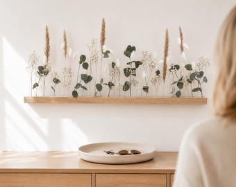 Puede incluir: Un estante rectangular de madera clara montado en la pared, con un arreglo de hojas de eucalipto secas, paniculata blanca y plumas de hierba de la pampa de color tostado. Debajo del estante hay una bandeja redonda de cerámica beige.