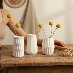 Könnte beinhalten: Drei weiße Keramikvasen mit geometrischen Mustern stehen auf einem Holztisch. Jede Vase enthält einen Stiel mit einer gelben, kugelförmigen Blume. Die Vasen variieren in Form und Größe und schaffen eine minimalistische Ästhetik.