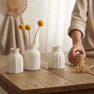 Könnte beinhalten: Drei kleine, weiße Keramikvasen mit unterschiedlichen Designs stehen auf einem Holztisch. Eine Vase enthält gelbe florale Akzente. Die Hand einer Person greift nach einem Holzobjekt. Die Vasen sind ein neutraler Einrichtungsgegenstand.