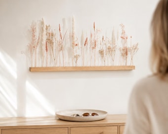Puede incluir: Una base de madera de tono claro y forma rectangular sostiene un arreglo de hierbas y flores secas en tonos crema, beige y rosa suave. Los tallos están colocados verticalmente en fila, creando una exhibición lineal de texturas variadas.