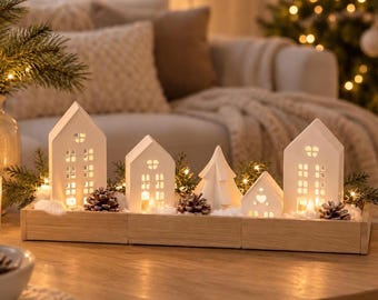 May include: A set of white ceramic house-shaped tea light holders and a tree figurine arranged on a rectangular wooden tray. The structures feature cut-out windows and doors, illuminated from within, surrounded by pine cones and faux snow.