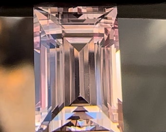 May include: A rectangular, faceted gemstone with a pale pink hue. The gem is held in a black setting, showcasing its geometric cuts and internal reflections. The stone's facets create a play of light and shadow.