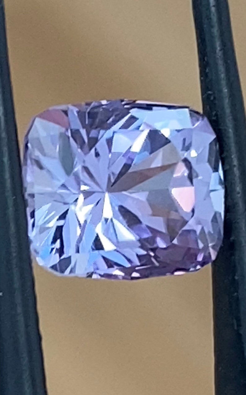 Op de afbeelding: Een lichtpaarse edelsteen met een kussen-slijpsel en vele facetten. De edelsteen wordt vastgehouden door een pincet.