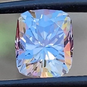Op de afbeelding: Een heldere, gefacetteerde edelsteen met een lichtblauwe tint en een regenboogkleurige schittering in roze en geel. De edelsteen is geslepen in een rechthoekige vorm met afgeronde hoeken.
