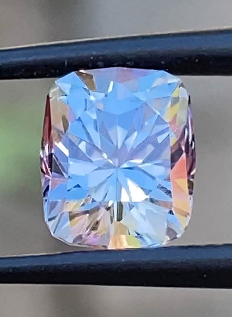 Op de afbeelding: Een heldere, gefacetteerde edelsteen met een lichtblauwe tint en een regenboogkleurige schittering in roze en geel. De edelsteen is geslepen in een rechthoekige vorm met afgeronde hoeken.