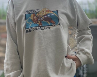 May include: A beige long-sleeve sweatshirt with an embroidered image of two anime characters fighting. The text "鬼滅の刃" and "あがきませんいっ" is also embroidered on the sweatshirt.