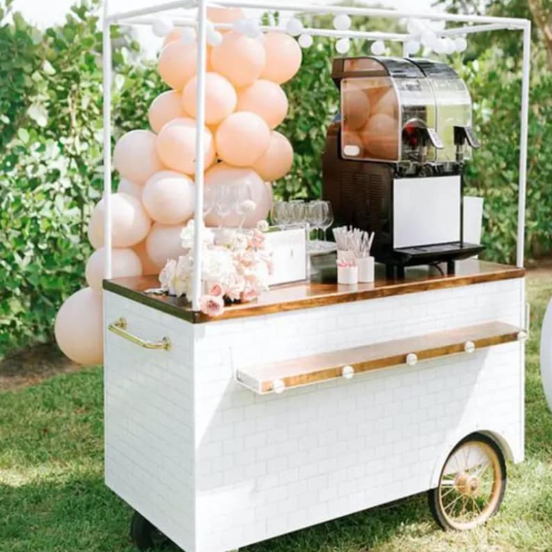 May include: A white food cart with a white brick pattern, a wooden countertop, and a gold handle. The cart has a black slushie machine and a display of peach-colored balloons.