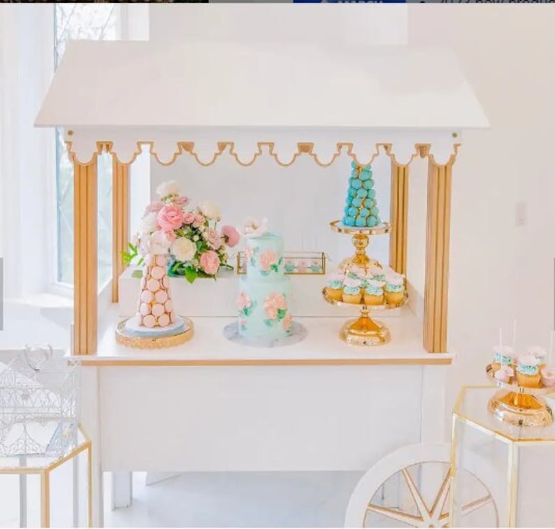 May include: White dessert cart with a gold trim and a white roof. The cart is decorated with pastel blue and pink macarons, cupcakes, and a cake. The cart is on a white surface.