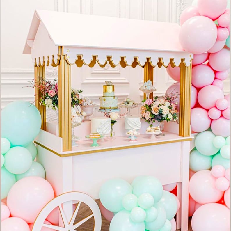 May include: A white wooden dessert cart with gold accents and a pink countertop. The cart is decorated with pastel pink and blue balloons and flowers. There are several cakes and pastries on display on the cart.