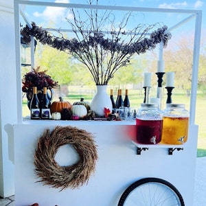 May include: White bar cart with a white metal frame and a white wooden base. The cart has a wreath on the side, two glass jars with spouts, and a variety of fall decorations, including pumpkins, gourds, and branches.