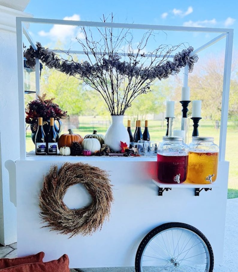 May include: White bar cart with a white metal frame and a white wooden base. The cart has a wreath on the side, two glass jars with spouts, and a variety of fall decorations, including pumpkins, gourds, and branches.