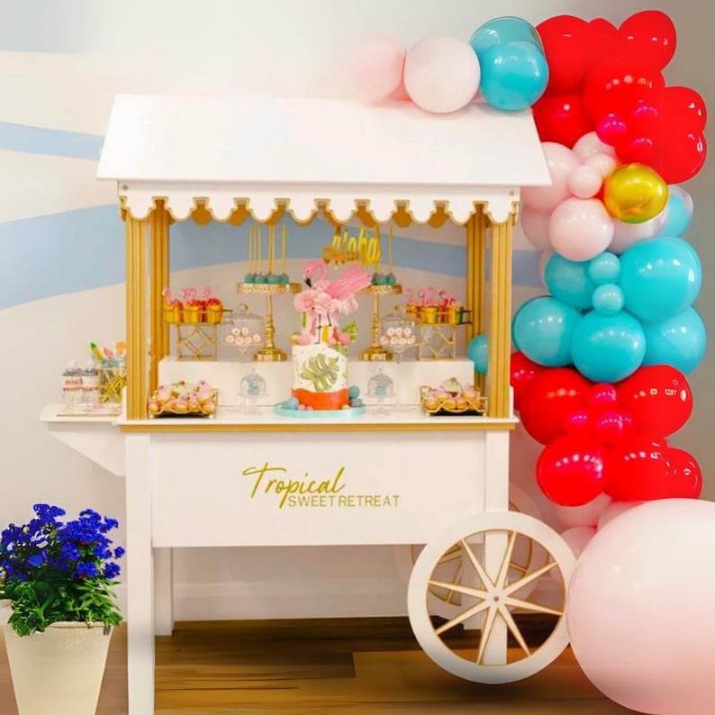 May include: A white dessert cart with gold accents and a sign that reads "Tropical Sweet Retreat". The cart is decorated with a pink flamingo cake, colorful candies, and a balloon arch in red, blue, and pink.