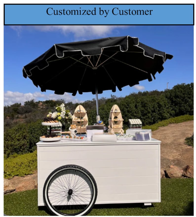 May include: White food cart with a black and white striped awning, a black umbrella, and a variety of desserts on display. The cart has a white wooden countertop and a large black wheel.