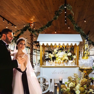 May include: A bride and groom dance in front of a white wedding cart decorated with flowers and gold accents. The cart has a sign that says "Love is Sweet".