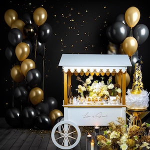 May include: A white dessert cart with gold accents and a sign that says "Love is Sweet". The cart is decorated with black and gold balloons, flowers, and candles.