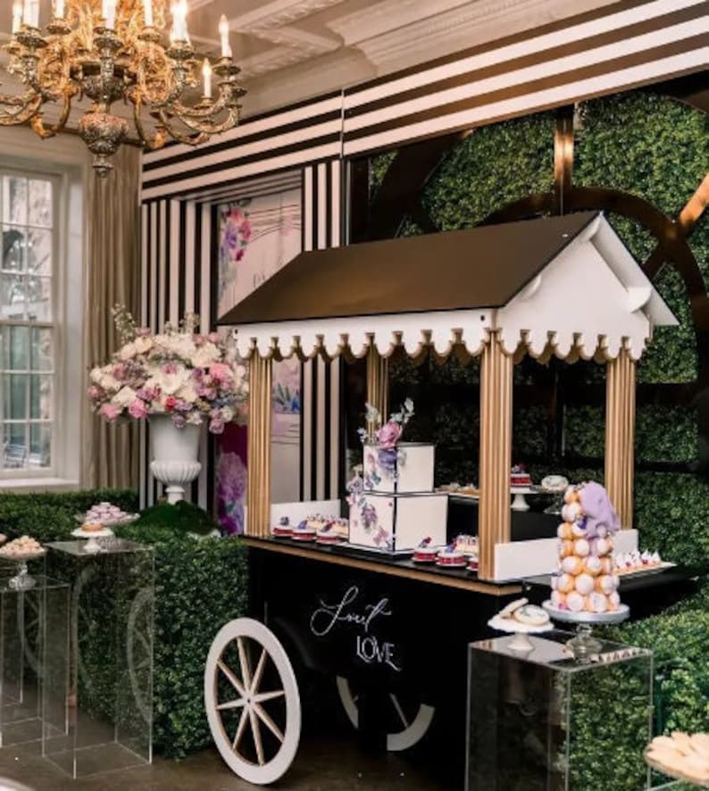 May include: A black and white striped backdrop with greenery behind a white and gold dessert cart with a black sign that says "Sweet Love". The cart is decorated with flowers and has a variety of desserts on display.