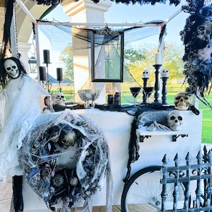 May include: A white Halloween-themed cart decorated with black spiderwebs, a spooky wreath, and a black metal gate. The cart has a black and white ghost, skulls, and candles on top.
