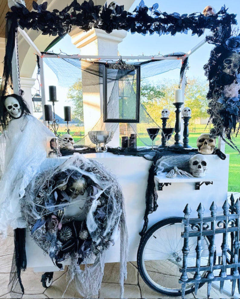 May include: A white Halloween-themed cart decorated with black spiderwebs, a spooky wreath, and a black metal gate. The cart has a black and white ghost, skulls, and candles on top.