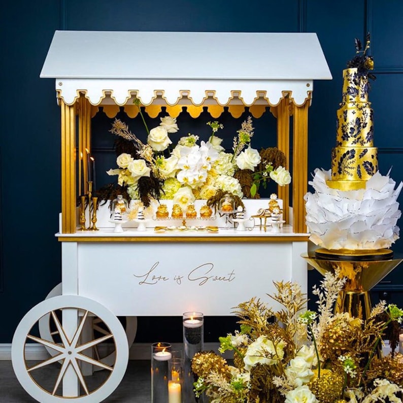 May include: A white dessert cart with gold accents and a sign that says "Love is Sweet". The cart is decorated with white flowers and a gold and black tiered cake.