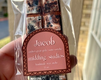 Puede incluir: Una barra de cera fundida marrón y negra con una etiqueta que dice "Jacob amber-pear-palo santo-smoke midding studios handmade wax melts 2 oz."