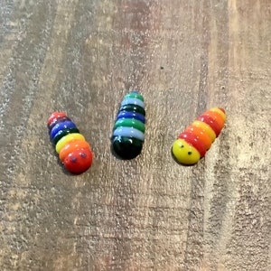 May include: Three small glass figurines shaped like caterpillars with colorful stripes. The caterpillars are red, orange, yellow, green, blue, and purple. They have black eyes and a yellow smile.