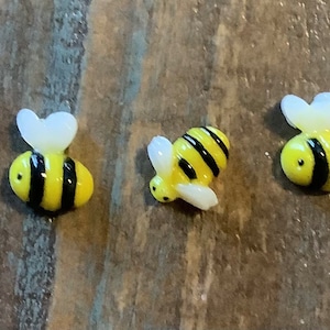 May include: Three small glass bee figurines with black and yellow stripes and white wings. The bees are arranged in a row on a wooden surface.