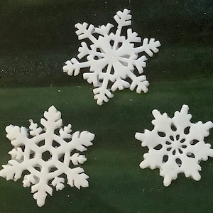 May include: Three white paper snowflakes on a dark green background. The snowflakes are different sizes and have intricate designs.
