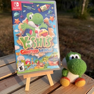 May include: A Nintendo Switch game case for Yoshi's Crafted World featuring a green Yoshi character on the cover, displayed on a small wooden easel next to a small, green, knitted yarn Yoshi plush toy with orange feet and a white belly.