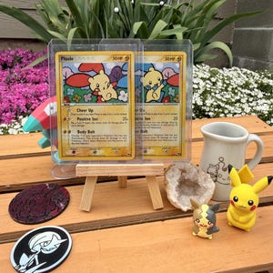 Könnte beinhalten: Zwei Pokemon-Sammelkarten mit dem Charakter Plusle, präsentiert in Schutzhüllen auf einer kleinen Holzstaffelei. Das Bild zeigt auch Pikachu-Figuren, eine Tasse und Untersetzer.