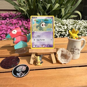Puede incluir: Una colección de artículos con temática Pokémon sobre una superficie de madera. Incluye una carta coleccionable Duskull, una figura de Porygon, una taza de Pikachu, una pequeña figura de Pikachu, dos pines redondos y una geoda. La carta se muestra en un caballete de madera en miniatura.