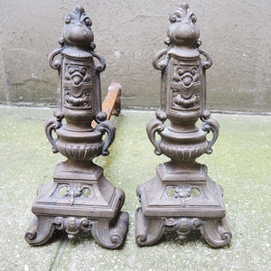 Puede incluir: Un par de fuegos de chimenea de latón antiguo con diseños ornamentados. Los fuegos de chimenea tienen una base decorativa con un motivo floral central y una columna alta y delgada con una cara estilizada en la parte superior.