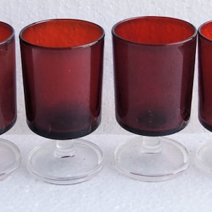 May include: Six clear stemmed glasses with a dark red hue. The glasses are all the same size and shape, and they are arranged in a row on a white surface.