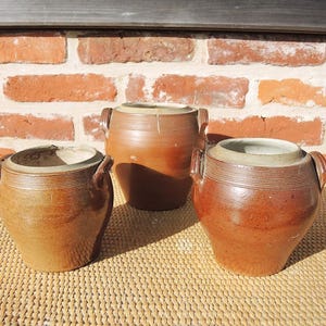 May include: Three brown glazed ceramic pots with two side handles each. The pots feature a rounded body, a wide opening, and horizontal ribbed detailing around the neck. The glaze varies in shade from light tan to deep reddish-brown.