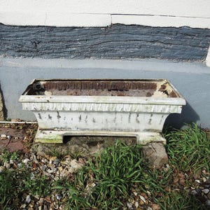 Può includere: Una fioriera rettangolare bianca e usurata con finiture decorative. La fioriera poggia su una base di pietra, circondata da erba e piccoli sassi. Il bordo superiore mostra segni di usura.