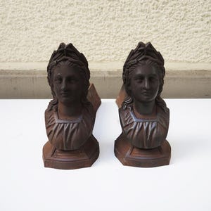 Peut inclure: Deux sculptures antiques en fonte de couleur marron foncé représentant des bustes de femmes. Chaque buste présente un visage détaillé, une couronne de laurier et un col carré. Les sculptures sont montées sur une base rectangulaire.